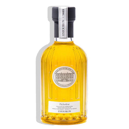 A bottle of Enskribos Picholine liqueur, featuring a ribbed clear glass bottle filled with golden yellow liquid, a white label with black text, and a black cap, set against a plain white background with a soft shadow.